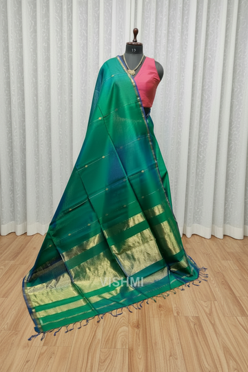 Green saree with gold border on a wooden floor with mannequin and other sarees in the background.